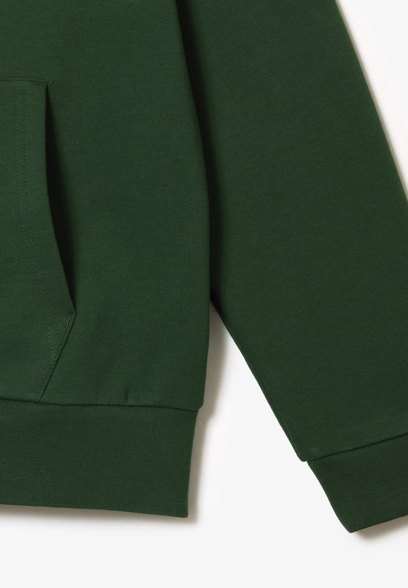 Manche et poche de sweat-shirt vert foncé avec poignets et ourlet côtelés sur fond blanc.