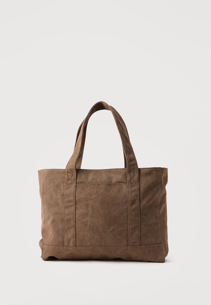 Sac fourre-tout en toile marron avec deux poignées et une poche avant, debout sur un fond clair uni.