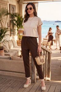 Donna con occhiali da sole che tiene un caffè freddo e una borsa di vimini, indossa una maglia beige e pantaloni marroni, in piedi su un pontile in legno accanto a un lounge sul mare.