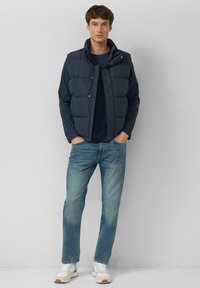 Marineblauw gewatteerd vest met drukknopen, gedragen over een donker shirt. Gecombineerd met lichtblauwe spijkerbroek en witte sneakers met beige accenten.