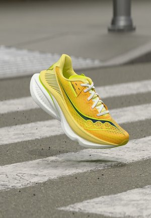 Chaussure de course jaune avec des lacets blancs et une semelle blanche épaisse en suspens au-dessus d'un passage piéton sur un asphalt gris.