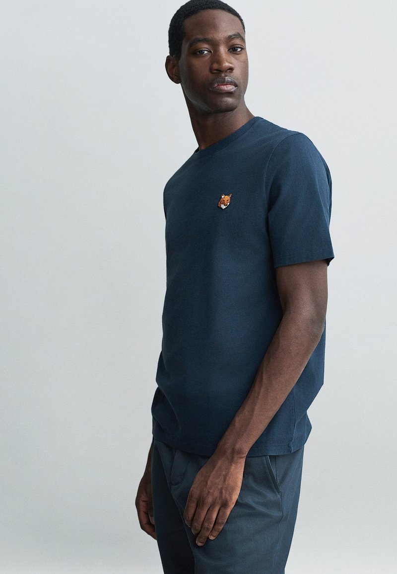 Tricou din bumbac de culoare albastru marin, cu mâneci scurte, guler rotund și un mic patch brodat în formă de vulpe pe partea stângă a pieptului. Textură netedă.