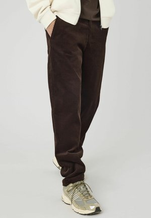 Pantalons en velours côtelé marron avec une texture douce, coupe fuselée et poches latérales. Associés à des baskets grises texturées.