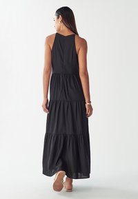Žena s dlouhými tmavými vlasy, oblečená v černých bezramínkových maxi šatech s volány a sandálech, stojící a otočená zády na bílém pozadí.