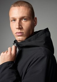 Ung mand med kort hår, der justerer kraven på en sort hættejakke, mens han ser direkte ind i kameraet mod en ensfarvet grå baggrund.