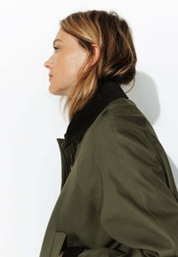Veste bomber verte avec un col en velours côtelé noir ; elle présente une fermeture éclair et une coupe décontractée. Texture lisse et détails de couture minimalistes.