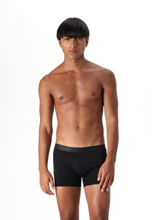 Schwarze Boxershorts aus dehnbarem Stoff mit glatter Textur. Der Bund hat ein dünnes, elastisches Band. Das Modell steht aufrecht, ohne Shirt.