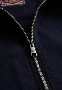 Gros plan sur la fermeture éclair d'une veste marine avec "SUPER DRY" gravé sur la languette métallique et une partie d'une étiquette de vêtement visible à l'intérieur.