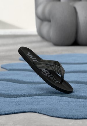 Tongs noires avec sangle tissée portant le logo « Wrangler », posées sur un tapis bleu texturé, avec un coussin gris flou en arrière-plan.