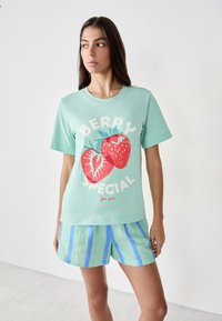 T-shirt verde menta con una grafica di fragola e testo bianco. Abbinata a pantaloni corti a righe nei toni del verde chiaro e blu.