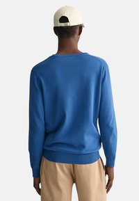 Blauer Strickpullover mit langen Ärmeln und einem gerippten Halsausschnitt, kombiniert mit hellbeigen Hosen, von hinten betrachtet. Mütze in Cremefarbe.