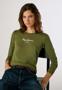 Olivgrünes Langarmhemd mit weißem "Pepe Jeans London"-Logo, kombiniert mit dunkelblauen Jeans, sitzend gegen einen schlichten schwarzen Stuhl.