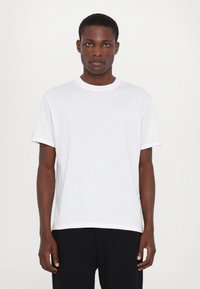 Tricou alb din bumbac, cu guler rotund, mâneci scurte și cusături contrastante portocalii la marginea mânecii și la marginea de jos. Prezentă o imprimare subtilă a logo-ului.