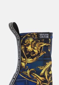 Botte en tissu à motifs bleus et noirs ornée de motifs décoratifs dorés, d'une surface texturée et d'une étiquette logo sur le côté.