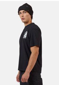 Černé bavlněné tričko s krátkým rukávem a kulatým výstřihem, na hrudi s bílou grafikou hory a logem. Muž nosí černou pletenou čepici.