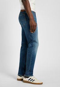 Blå denimjeans med rak skärning, medelhög midja, urblekt finish och femfickdesign, tillsammans med krämfärgade sportskor.