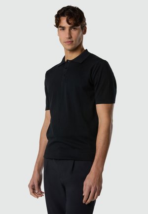 Junger Mann mit lockigem Haar, der ein schwarzes Poloshirt mit kurzen Ärmeln und schwarze Hosen trägt, vor einem hellgrauen Hintergrund stehend.