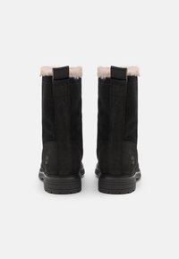 Botas de tobillo de ante negro con un suave forro de piel de oveja beige en la parte superior y una suela de goma. Presenta un lazo en la parte posterior para facilitar el uso.
