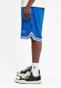 Pantalones cortos de baloncesto azules con rayas blancas, bolsillo lateral y logo cerca del dobladillo. Zapatillas negras con cordones blancos y suelas texturizadas. Calcetines blancos visibles.