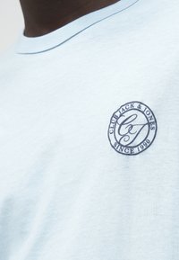 Camiseta de manga corta azul claro hecha de un tejido suave, con un logo redondo azul marino que dice "Club Jack & Jones Desde 1990" en el pecho.