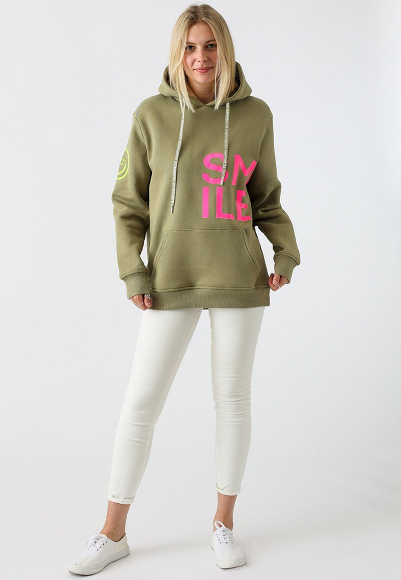 Olijf groene hoodie met roze "SMILE" print, kangoeroe zak, trekkoordsluiting en geribbelde manchetten, gecombineerd met witte broek en sneakers.