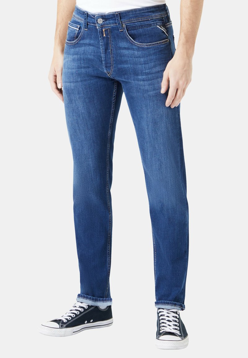 Sötétkék farmernadrág, slim fit fazonban, öt zsebbel és kontrasztos varrással. Fekete-fehér alacsony szárú tornacipővel viselve.