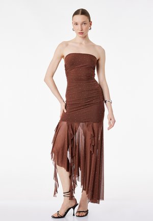 Kvinde iført en stropløs brun kjole med små rhinsten, en asymmetrisk gennemsigtig flæsekant, sorte højhælede sandaler og sølvfarvede creoløreringe.