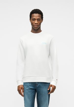 Mężczyzna w białej bluzie z subtelnym logo na piersi i rękawie, w średnio niebieskich dżinsach, stoi na tle jednolitego białego tła.