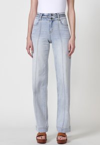 Jeans taille haute bleu clair avec une ceinture tressée, coupe droite et lignes de couture visibles. Portés avec des sandales brunes à lanières.
