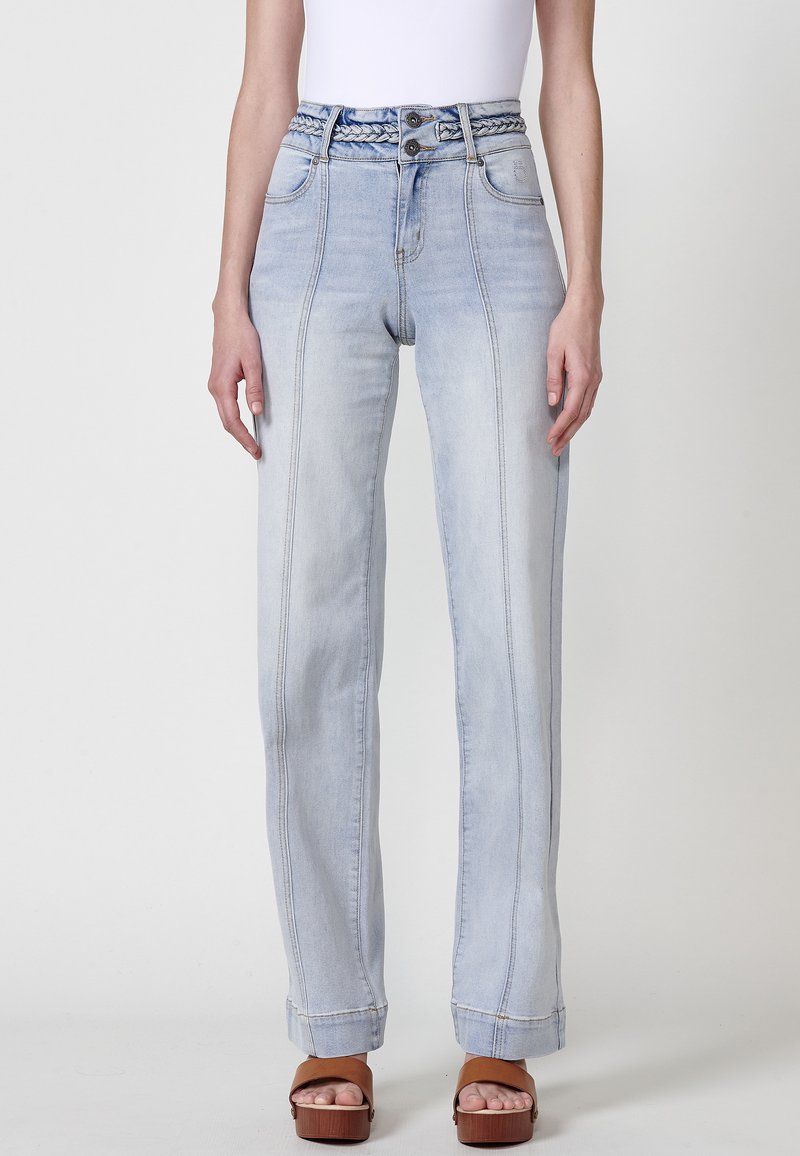 Jeans taille haute bleu clair avec une ceinture tressée, coupe droite et lignes de couture visibles. Portés avec des sandales brunes à lanières.
