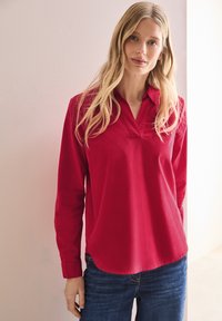 Chemise rouge à manches longues avec un col cranté, en tissu lisse. Ourlet arrondi, silhouette ajustée, portée avec un jean en denim bleu.