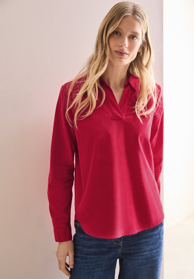 Chemise rouge à manches longues avec un col cranté, en tissu lisse. Ourlet arrondi, silhouette ajustée, portée avec un jean en denim bleu.