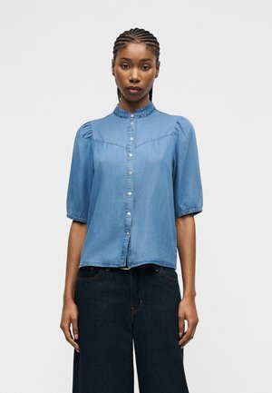 Kobieta z zaplecionymi włosami, ubrana w jasnobłękitną koszulę na guziki z bufkami na rękawach oraz ciemne, szerokie jeansy.