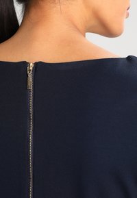 Granatowa sukienka o minimalistycznym designie, z widocznym zapięciem na zamek z tyłu i gładkim, teksturowanym materiałem.