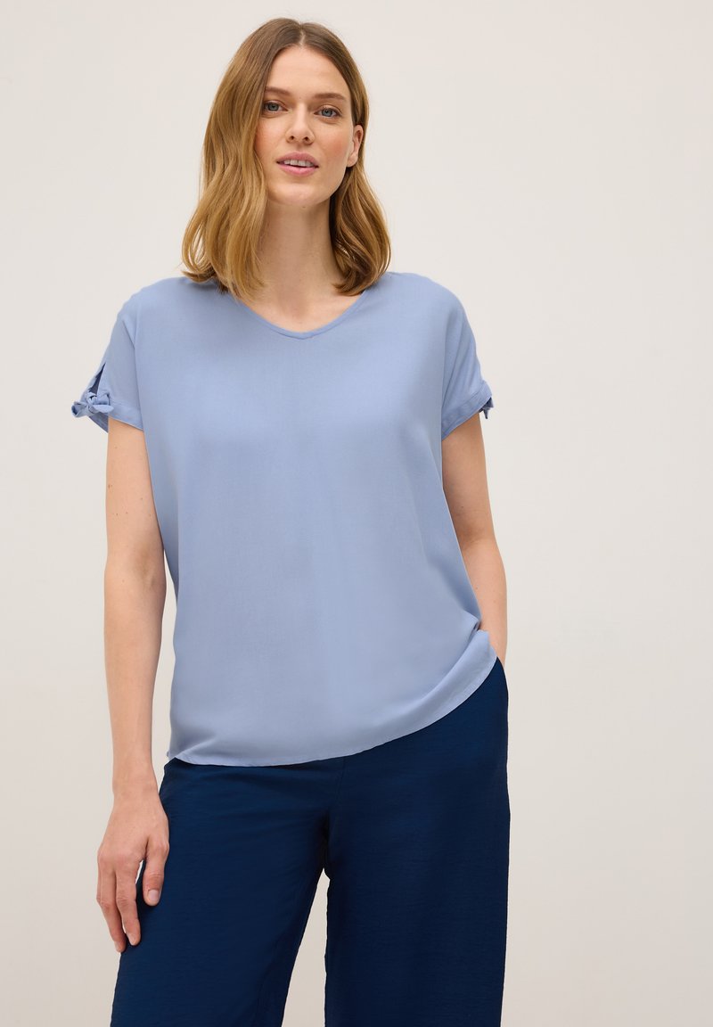 Lichtblauwe blouse met korte mouwen, een v-hals en geknoopte mouwdetails, gemaakt van zacht materiaal, gecombineerd met marineblauwe wijde broek.