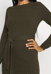 Mörk olivgrön ribbad stickad klänning med långa ärmar och matchande bälte knutet i midjan, hand vilande på höften.