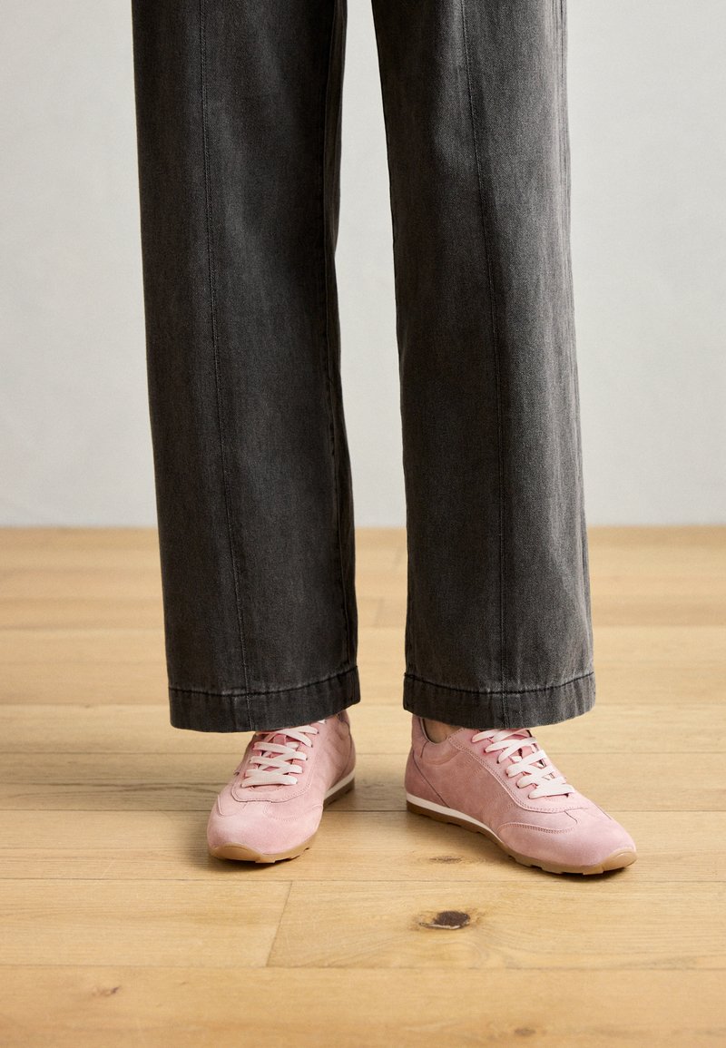 Roze suède sneakers met witte veters, gecombineerd met brede, grijze broeken. De houten vloer op de achtergrond benadrukt het ontwerp van het schoeisel.