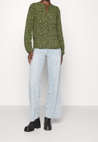 Blouse noire avec un motif floral vert, à manches longues et décolleté en forme de clé. Associée à un jean large bleu clair et des bottes à plateforme noires.
