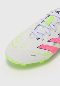 Botas de fútbol blancas con acentos en rosa vibrante y verde neón, parte superior sintética texturizada y cierre tradicional con cordones. Presenta la etiqueta "HYBRIDFEEL".