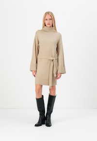 Beige stickad klänning med hög krage, långa ärmar och knytbart midjeband. Har ribbade detaljer vid ärmsluten och nederkanten. Bärs med svarta knähöga stövlar.