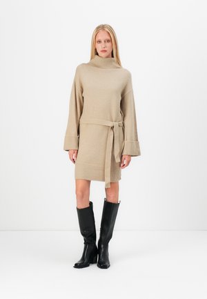 Beige stickad klänning med hög krage, långa ärmar och knytbart midjeband. Har ribbade detaljer vid ärmsluten och nederkanten. Bärs med svarta knähöga stövlar.