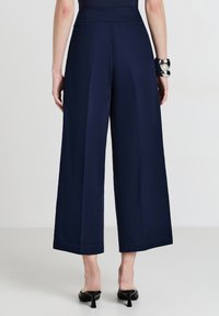 Donna che indossa pantaloni navy a vita alta e gamba larga, con mule nere con tacco, vista da dietro su uno sfondo neutro.