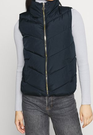 Femme portant une veste matelassée bleu marine avec une fermeture éclair dorée sur un haut côtelé à manches longues gris clair et un jean gris.