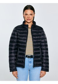 Sieviete, kas valkā melnu dūrienu žaketi virs strīpainas kreklas un gaiši zilas džinsu bikses, stāvot pret vienkāršu gaišu fonu.