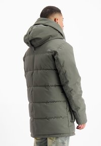 Man in een groene gewatteerde winterjas met een grote capuchon, gezien van achteren tegen een witte achtergrond.