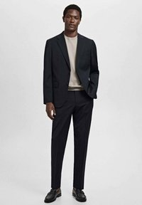Sacou negru, croi adaptat, cu rever în formă de ureche, purtat cu un tricou de culoare deschisă și pantaloni asortați. Pantofi din piele neagră completează ținuta.