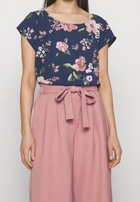 Floral-printed navy top with a round neckline and short sleeves, paired with light pink high-waisted trousers featuring a tie belt.