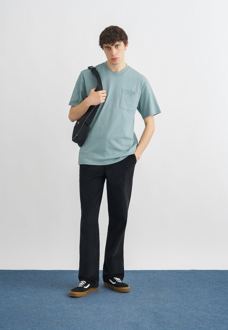 Un giovane uomo è in piedi su un tappeto blu indossando una t-shirt teal con taschino, pantaloni neri, sneakers nere e portando una borsa a tracolla nera.