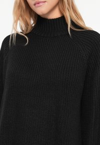 Melnas adītas džemperis ar augstu apkakli un ribošanu, brīvs piegrieziens un raglanu piedurknes.