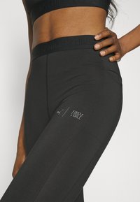 Leggings neri realizzati in materiale elasticizzato, con un cinturino testurizzato con la scritta "FIRST MILE" in rilievo e un logo stampato sulla coscia.